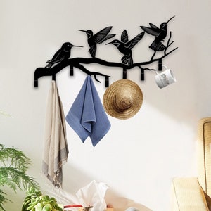 May include: Black metal wall-mounted coat rack shaped like a branch with hummingbirds perched on it. The rack has five hooks and is decorated with a straw hat and a blue towel hanging from the hooks.