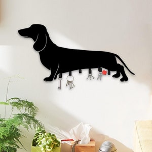 May include: Black metal dachshund silhouette wall mounted key holder with keys hanging from it. The dachshund is on a white wall with a plant, a pumpkin, and a tissue box in the foreground.