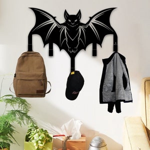 May include: Black metal bat wall hook with three hooks. The hooks are holding a brown backpack, a black baseball cap, and a gray jacket. The bat is silhouetted against a white wall.