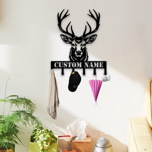 May include: Black metal deer head wall hook with the text "CUSTOM NAME" above it. The hook has six hooks for hanging items. A pink umbrella, a black baseball cap, and a brown scarf are hanging from the hooks.