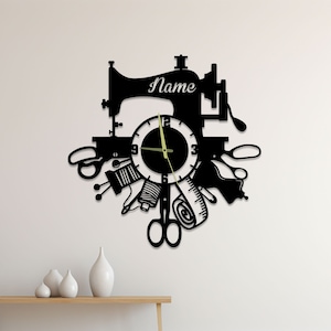 May include: Black wall clock featuring a sewing machine design. The clock face is encircled by sewing-related items such as scissors, thread, and a tape measure. The clock face has a black background with white numbers and gold hands. The sewing machine has the word "Name" written on it.