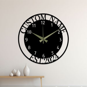 Può includere: Orologio da parete in metallo nero con numeri e lancette bianchi. Il quadrante dell'orologio ha il testo "CUSTOM NAME EST 2024" attorno al bordo.