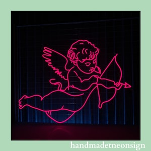 May include: Pink neon sign depicting a cupid with wings and an arrow. The sign is on a black background with a grid pattern.