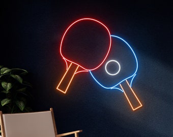 May include: Neon sign featuring two ping pong paddles, one red and one blue, with orange handles. The sign is mounted on a dark blue wall, with a wooden chair and plant in the foreground. The neon lights create a vibrant and modern aesthetic.