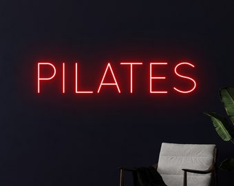 May include: Red neon sign with the word "PILATES" on a dark wall. A white chair with a black throw blanket is in the foreground.