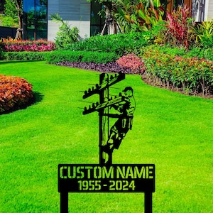 May include: Black metal silhouette of a lineman climbing a utility pole with power lines. The silhouette is mounted on a black metal stake with a sign that reads "CUSTOM NAME 1955 - 2024".