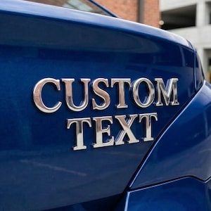 May include: A dark blue car boot lid featuring shiny, metallic, three-dimensional lettering that reads CUSTOM TEXT. The chrome-finished letters are individually applied to the smooth, glossy surface of the vehicle's rear exterior panel.
