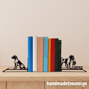 May include: Black metal bookends featuring a silhouette of a dog with the name "COOPER" on the right side. The bookends are holding up a row of colorful books.