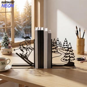 May include: A pair of black metal bookends featuring a silhouette design. One piece depicts Santa Claus skiing, while the other shows a forest scene with pine trees and a winding path. The bookends are holding four books upright on a wooden surface.