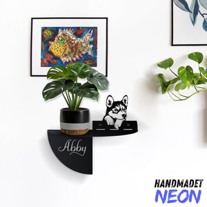 Op de afbeelding: Zwart metalen wandrek met een halfronde vorm. Het rek heeft een uitsnijding van een husky-hond en de naam "Abby" staat op het rek. Een plant in een pot staat op het rek.