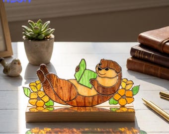 May include: A stained glass otter figurine, lying on its back and reading a green book. The otter is brown and tan, with yellow flowers and green leaves at its base. The figurine sits on a wooden base. The text "HANDMADET NEON" is in the upper left corner.