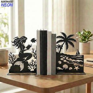 May include: Black metal bookends featuring a mermaid reading and a beach scene with a sailboat, palm trees, and seashells. The bookends are holding several books on a wooden table. The word "HANDMADET NEON" is in the top left corner.