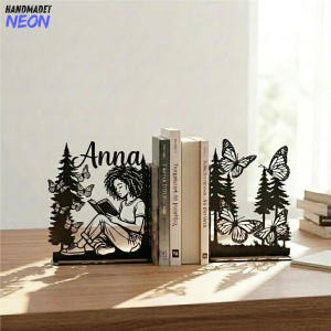 May include: A black metal bookend set featuring a silhouette of a person with curly hair reading a book under pine trees with butterflies. The left piece includes the name Anna in script. The right piece features pine trees and butterflies.