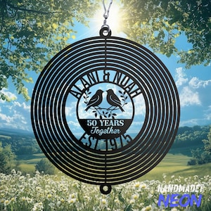 May include: A black metal wind spinner with a circular design. The center features the names "ALANI & NOAH", two birds, and the text "50 YEARS Together EST 1975". The spinner hangs from a silver hook, with a scenic background.