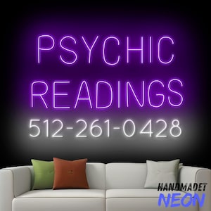 May include: Purple neon sign with the text "PSYCHIC READINGS" and the phone number "512-261-0428" in white neon. The sign is hanging on a wall behind a white couch with two pillows.