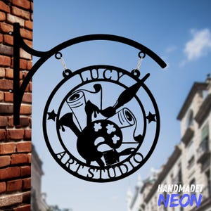 May include: Black metal sign with a circular design featuring a paintbrush, paint palette, and scrolls. The sign reads "Lucy Art Studio" and "Handmade Neon".