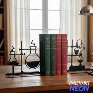 以下が含まれることがあります： 実験用フラスコのデザインの黒い金属製ブックエンド。緑と赤の表紙の本が並んでいます。ブックエンドは木製の机の上にあり、背景には窓とランプがあります。「HANDMADET NEON」の文字が見えます。
