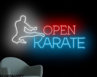 May include: A neon sign featuring a white silhouette of a person performing a karate kick next to the red text OPEN and the blue text KARATE. The sign is mounted on a dark wall above a light blue armchair, with the text HANDMADET NEON in blue.