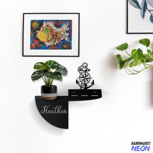 May include: Black metal shelf with a half-circle design. The shelf has a black anchor with a compass design and the word "Heather" written in white on the front. A potted plant sits on the shelf.
