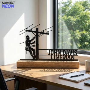May include: Black metal lineman silhouette on a wooden base. The silhouette depicts a lineman working on a power pole. The name "JOHN DAVIS LINEMAN" is cut out below the silhouette. The word "HANDMADET NEON" is in the upper left corner.
