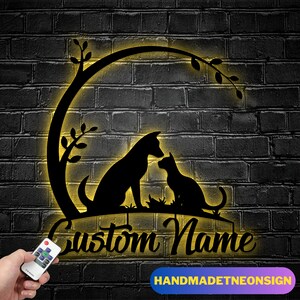 May include: Black metal sign featuring a dog and cat silhouette under a tree branch, with the words "Custom Name" in a cursive font. The sign is illuminated with a warm yellow glow against a dark brick wall. The sign also has the text "HANDMADENEONSIGN".