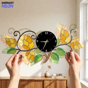 May include: A horizontal wall clock featuring a central black circular face with white numbers and gold hands, flanked by stained glass elements in yellow and green hues shaped like hop cones and leaves, framed in dark metal scrollwork.