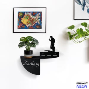 Op de afbeelding: Zwart metalen wandrek met een silhouet van een persoon die een vergrootglas vasthoudt. Het rek is gebogen en heeft de naam "Zachary" erop geschreven in witte letters. Een plant in een pot staat op het rek.