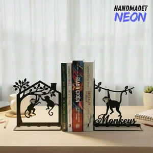 May include: Black metal bookends featuring monkey silhouettes. One bookend depicts monkeys in a tree and house, the other shows a monkey hanging from a branch. The word "Monkeys" is written on the base of the second bookend. The text "HANDMADET NEON" is in the upper right corner.