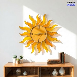 May include: A decorative sun-shaped wall clock with a yellow and orange stained glass design. The clock face displays the numbers 12, 3, 6, and 9. The clock is mounted on a white wall above a wooden cabinet. The text "HANDMADET NEON" is in the upper right corner.