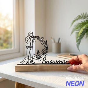 Pode incluir: Uma silhueta de metal preto de uma pessoa a cantar num microfone com notas musicais rodopiantes e uma clave de sol, apresentando a palavra "Snowman" em letra cursiva, montada numa base retangular de madeira de cor clara.
