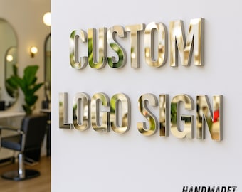 May include: A wall-mounted sign with the words "CUSTOM LOGO SIGN" in metallic gold letters. The sign is displayed on a white wall, with the text "HANDMADET NEON" in blue at the bottom.