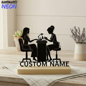 Puede incluir: Silueta negra de una manicurista y clienta en una mesa de salón, montada sobre una base de madera. Las palabras "CUSTOM NAME" están recortadas debajo de las figuras. El texto "HANDMADET NEON" está en la esquina superior izquierda.