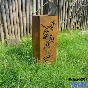 May include: A tall, rectangular, weathered metal planter with a pine cone and branch design cut into the sides. The planter is a rusty brown colour and sits in a bed of green grass. The text "HANDMADET NEON" is visible at the bottom.