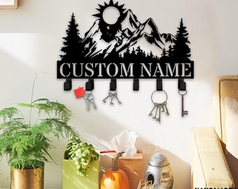 May include: A black metal wall-mounted key holder featuring a mountain range silhouette, a sun, and pine trees. The design includes the text CUSTOM NAME in capital letters and six hooks, with several metal keys hanging from the hooks.