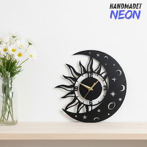 May include: Black wall clock in a sun and moon design. The clock face is white with gold hands and black numbers. The clock is shaped like a crescent moon with stars and circles. The text "HANDMADET NEON" is in the upper right corner.