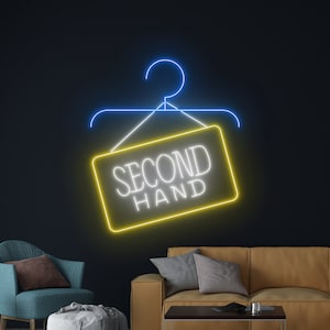 Puede incluir: Un letrero de neón con forma de percha con las palabras "SECOND HAND" en letras blancas sobre un rectángulo amarillo. La percha es azul y el letrero cuelga de una pared azul oscuro. Un sillón verde azulado y un sofá marrón claro están en el fondo.