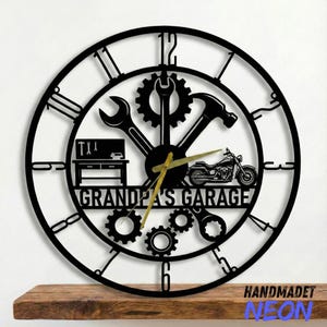 May include: Black metal wall clock with Roman numerals and gold hands. The clock features a motorcycle, tools, and the text "Grandpa's Garage". The clock is mounted on a wooden shelf.