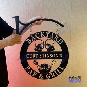 Może przedstawiać: Czarny metalowy szyld do baru i grilla na podwórku z napisem "BACKYARD BAR & GRILL" i "CURT STINSON'S". Szyld przedstawia kufle do piwa i grafikę grilla. Jest zawieszony na czarnym metalowym wsporniku.