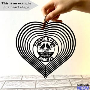 Puede incluir: Un m&oacute;vil e&oacute;lico en forma de coraz&oacute;n de metal negro con c&iacute;rculos conc&eacute;ntricos. El centro presenta el texto "Connie & Paul 50 Years Together EST. 1975" con dos p&aacute;jaros. Tambi&eacute;n est&aacute; presente el texto "This is an example of a heart shape".
