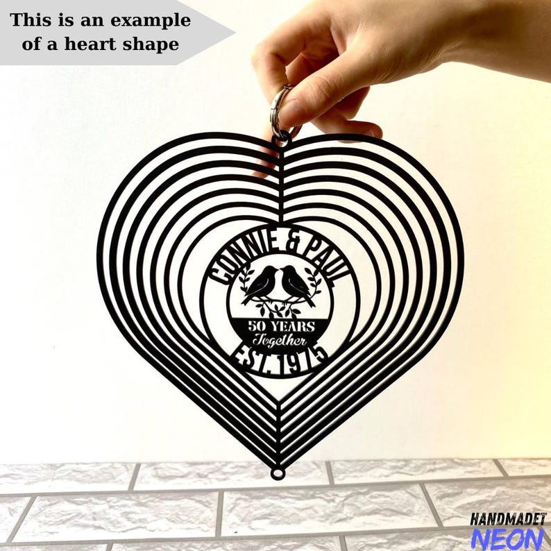 Puede incluir: Un m&oacute;vil e&oacute;lico en forma de coraz&oacute;n de metal negro con c&iacute;rculos conc&eacute;ntricos. El centro presenta el texto "Connie & Paul 50 Years Together EST. 1975" con dos p&aacute;jaros. Tambi&eacute;n est&aacute; presente el texto "This is an example of a heart shape".