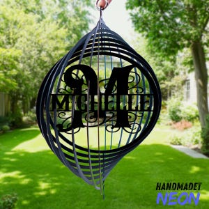May include: Black metal wind spinner with the name "Michelle" and the number "94" cut out. The wind spinner is suspended in front of a green lawn and trees. The text "HANDMADET NEON" is at the bottom.