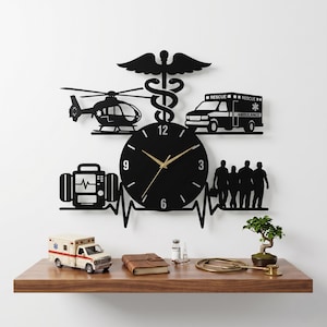 May include: Black metal wall clock with medical symbols, including a helicopter, ambulance, and caduceus. The clock face has gold hands and a heartbeat design. A miniature ambulance and other items sit on a wooden shelf below.