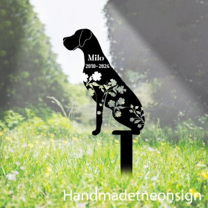 Puede incluir: Silueta de perro de metal negro con detalles florales blancos, una placa conmemorativa con el nombre "Milo" y las fechas "2010-2024", y una estaca de metal negro para uso en exteriores.