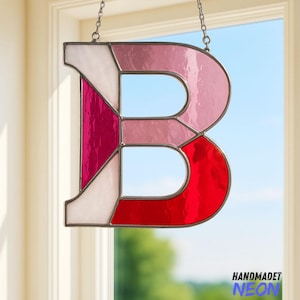 May include: A stained glass letter "B" in shades of pink, red, and white, hanging from a chain. The letter is segmented with metal borders. The word "HANDMADET NEON" is written in blue and purple at the bottom right.
