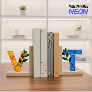 May include: A decorative bookend set featuring the letters "V" and "T" in stained glass. The "V" is yellow and orange, and the "T" is blue. Each letter has a green leaf accent. The bookends are on a wooden base, with books in between.