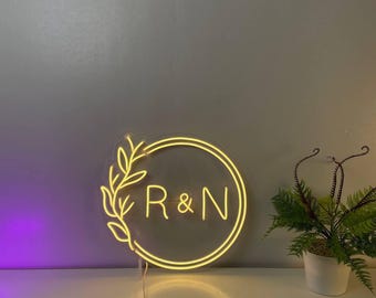 May include: A circular neon sign emitting a warm yellow light, featuring the letters R & N centered within a double-ring border. A stylized leafy branch design extends along the left side of the circle. The sign is mounted on a clear acrylic backing.