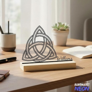 Puede incluir: Un diseño de nudo celta negro sobre una base de madera, con el nombre "Willams" grabado en la base. El diseño es un símbolo de interconexión. La base es rectangular y de madera clara. El fondo incluye un escritorio, un portalápices y una planta suculenta.