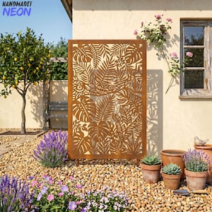 Puede incluir: Una pantalla metálica rectangular de color óxido con un diseño de hojas tropicales cortadas con láser. La pantalla está al aire libre, en un jardín con un lecho de grava, plantas en macetas y un limonero. El texto "HANDMADET NEON" es visible.