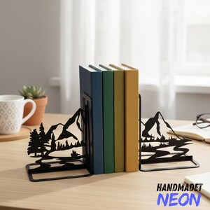 May include: Black metal mountain-themed bookends holding colorful books. The bookends feature a mountain and tree silhouette design. The books are blue, green, and yellow. The bookends are on a wooden surface.