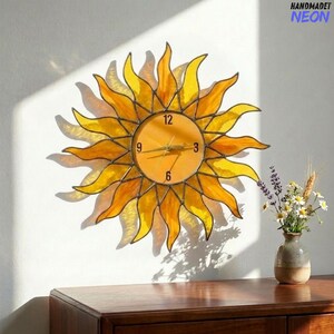 May include: A handcrafted sunburst clock with a yellow and orange stained glass design. The clock face displays the numbers 12, 3, 6, and 9. The clock is mounted on a white wall. The text "HANDMADET NEON" is in the upper right corner.